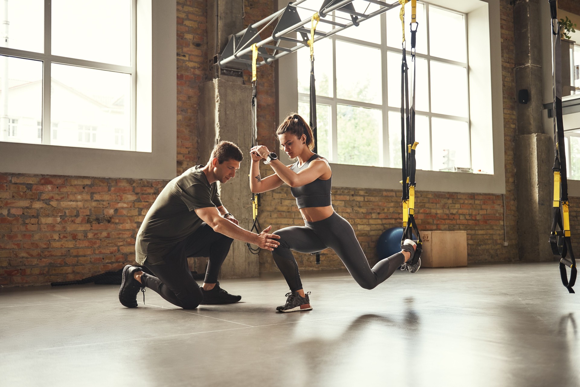 Doing squat exercise. Confident young personal trainer is showing slim athletic woman how to do squats with Trx fitness straps while training at gym.